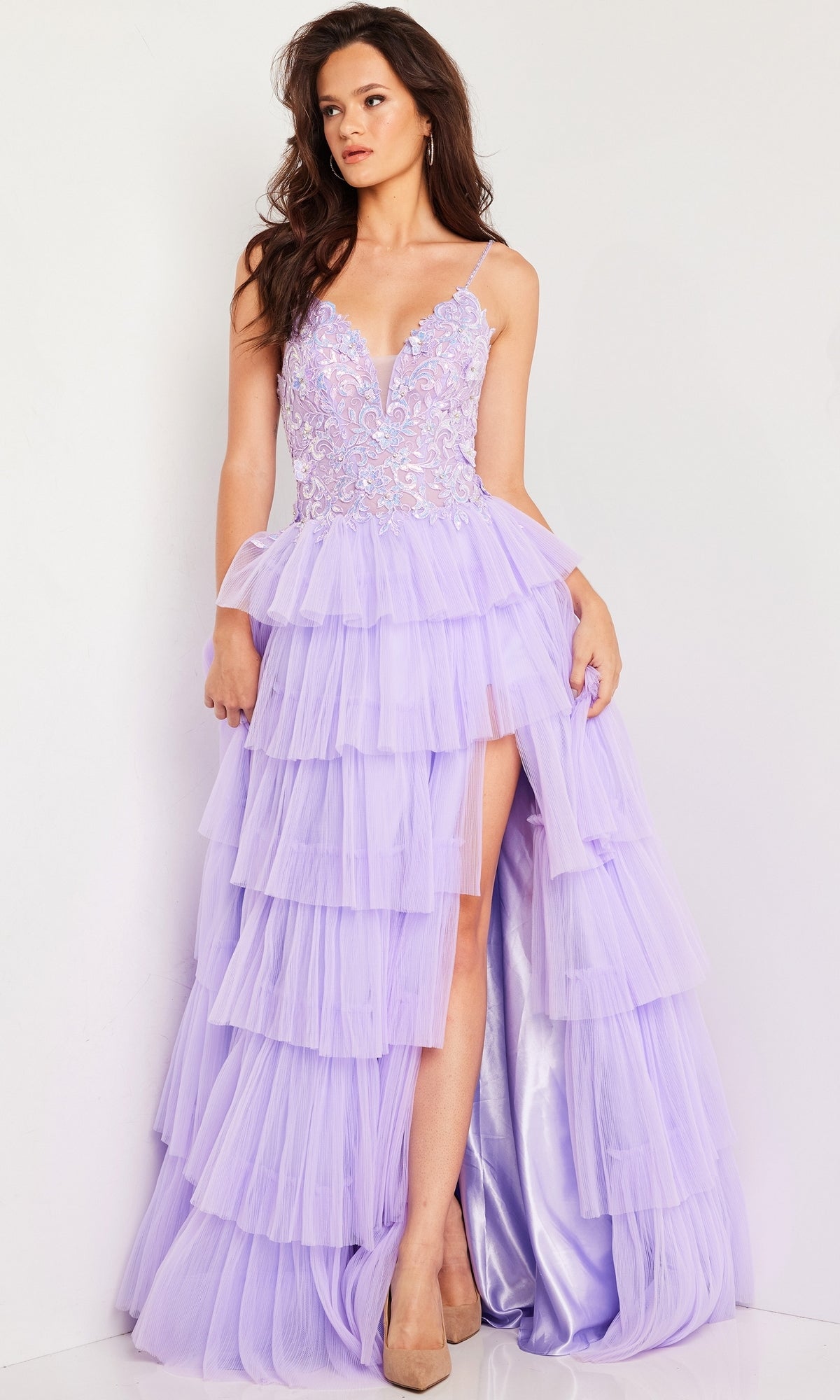 A woman stands against a plain white background wearing the Long Prom Dress 37190 by Jovani— a light purple tulle ball gown with ruffles, a lace embellished bodice, and a high slit.